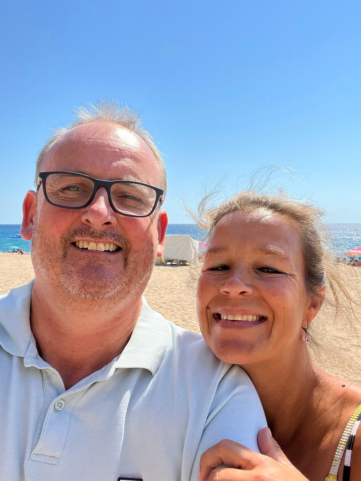 Een glimlachend stel geniet van de zon op het zandstrand van L'Estartit. Man met bril, vrouw met blond haar, beiden dragen zomerkleding.